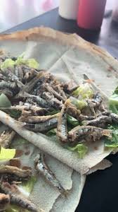 Flatbread topped with small fried fish and green vegetables, this Sardine Sandwich is served on a dark surface with sauce bottles blurred in the background.